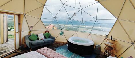 Une pièce en forme de dôme géodésique comprenant un lit, un canapé, un jacuzzi et offrant une vue sur le paysage.