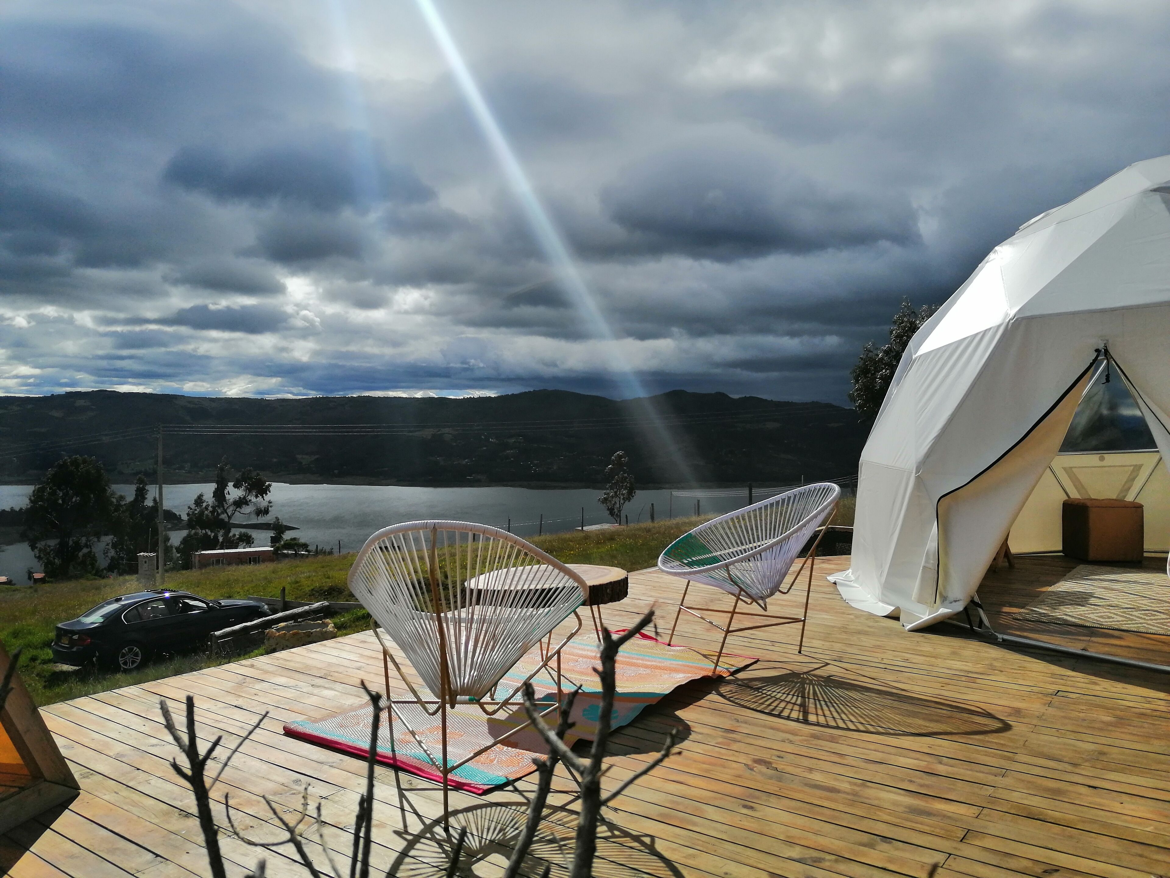 A tented accommodation with a wooden deck, two white chairs, and a view of a lake and mountains.