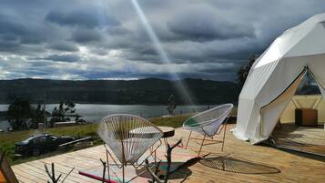A tented accommodation with a wooden deck, two white chairs, and a view of a lake and mountains.
