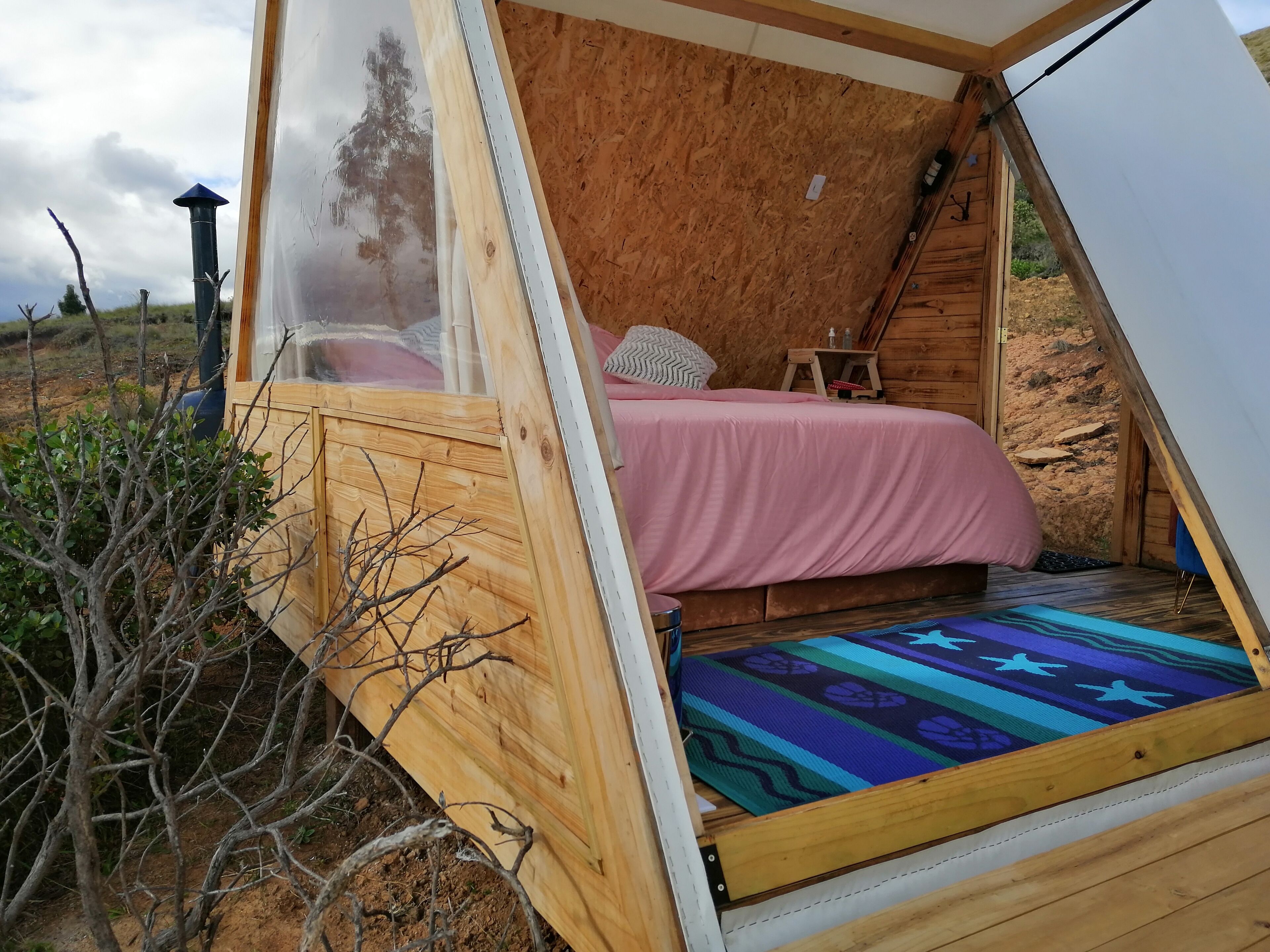 Una tienda de madera triangular acogedora, con una cama, almohadas y una mesita.