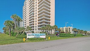 A high-rise building with multiple balconies, a sign for 'SANTA ROSA DUNES,' and a road in the foreground. - 🏝️Sound Views | Walk to Beach☀️ Santa Rosa Dunes 722 (Pensacola Beach)