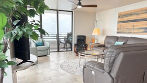 A modern living room with a sofa, armchairs, a coffee table, and a TV. There is a ceiling fan, a lamp, and a painting on the wall. - Oceanfront Condo Wraparound Balcony Sunrise Views (Daytona Beach)