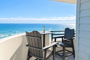 A balcony with two wooden chairs and a table, overlooking a beach with the ocean and a clear sky. - Oceanfront Condo Wraparound Balcony Sunrise Views (Daytona Beach)