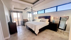 A spacious bedroom with a large bed, a ceiling fan, a desk, and a view of the outdoors through the windows. - Oceanfront Condo Wraparound Balcony Sunrise Views (Daytona Beach)