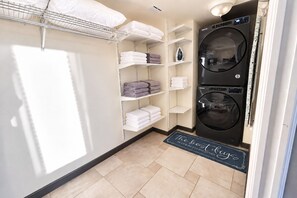 A laundry room with a Samsung washer and dryer, shelves with folded towels, and a door mat with the text 'The bed in life, clean clothes.' - Oceanfront Condo Wraparound Balcony Sunrise Views (Daytona Beach)