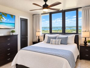 A hotel room with a bed, a ceiling fan, a TV, a window with a view, and a dark wooden dresser.