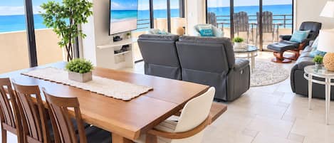 A modern living room with a wooden dining table, grey sofas, and a view of the ocean.