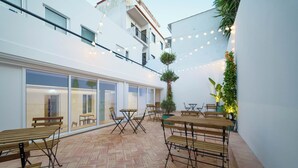 Un patio al aire libre con mesas y sillas de madera, piso con patrón de espiga y luces colgantes.