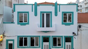 Un edificio de dos pisos con fachada blanca y detalles en color verde azulado. Cuenta con varias ventanas, un balcón y una base de ladrillo.