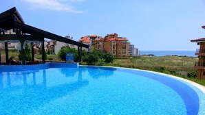 A swimming pool with a clear blue water surface, surrounded by a covered area with a roof, and buildings in the background. - Menada Paradise Dreams Apartments (Sveti Vlas)
