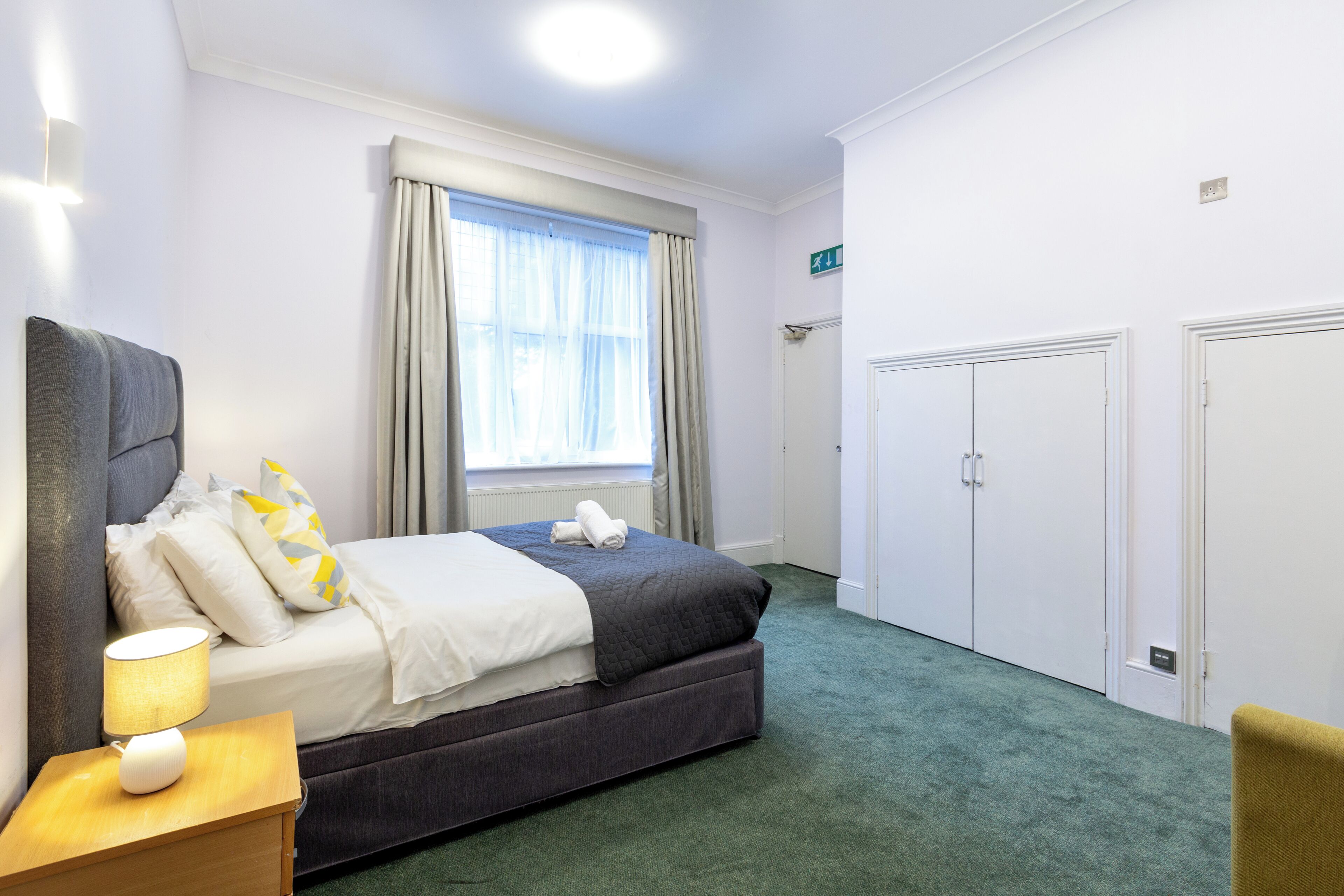 A hotel room with a bed, a nightstand, a window with curtains, a wardrobe, and a door.
