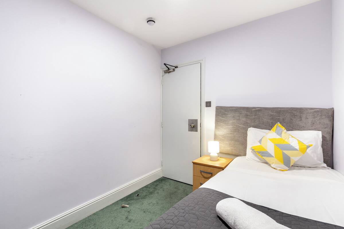 A bedroom with a bed, a bedside table with a lamp, a door, and a carpeted floor.