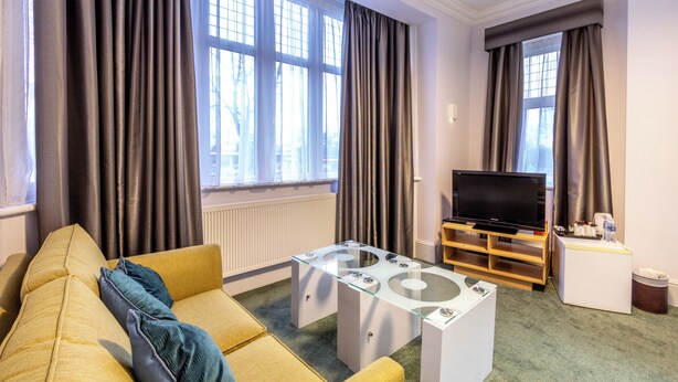 A living room with a yellow sofa, a glass coffee table, a television on a wooden stand, and large windows with curtains. - London Gatwick Inn (Horley)