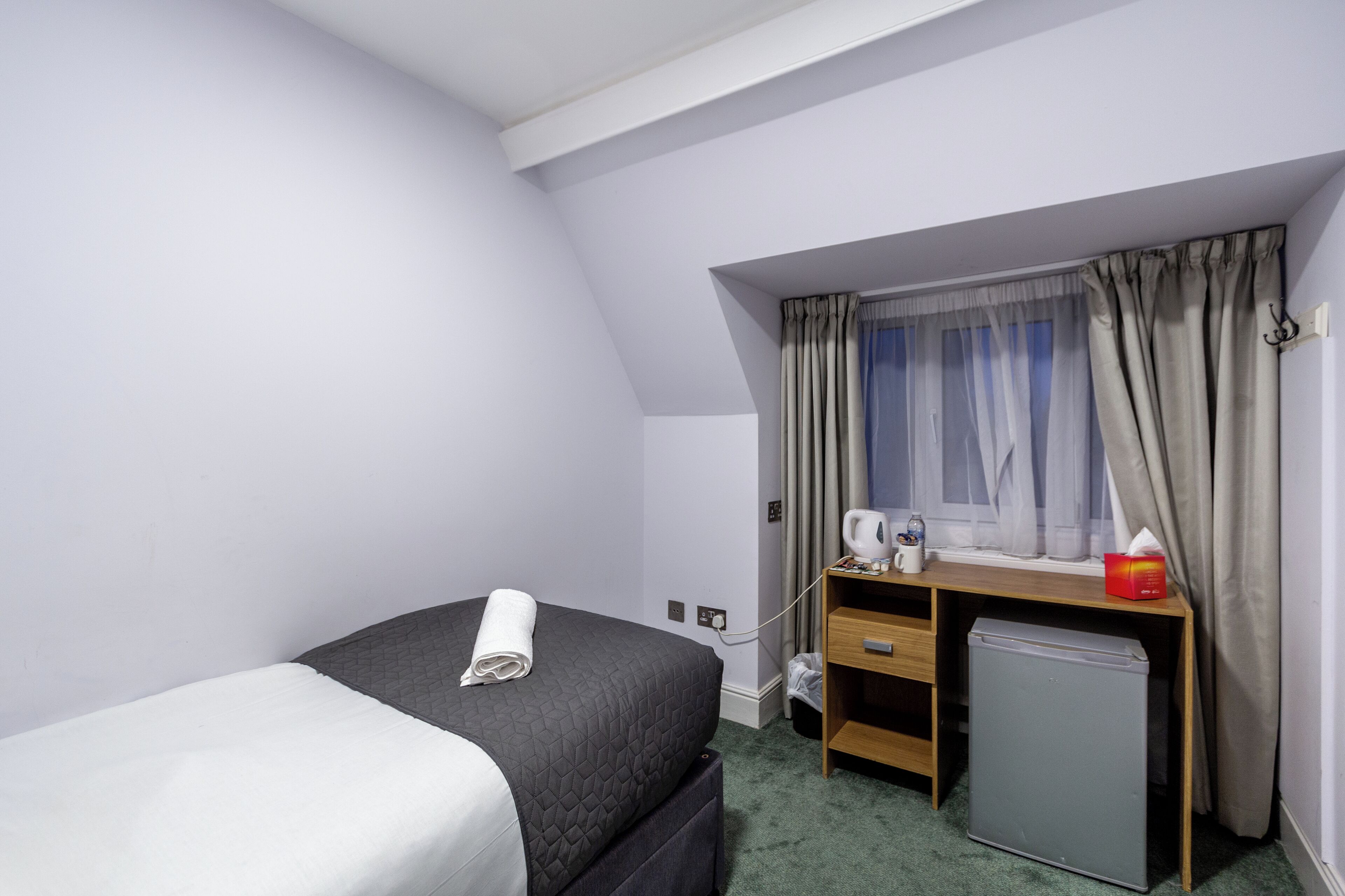A hotel room with a bed, a desk with a computer, a small fridge, and a window with curtains.
