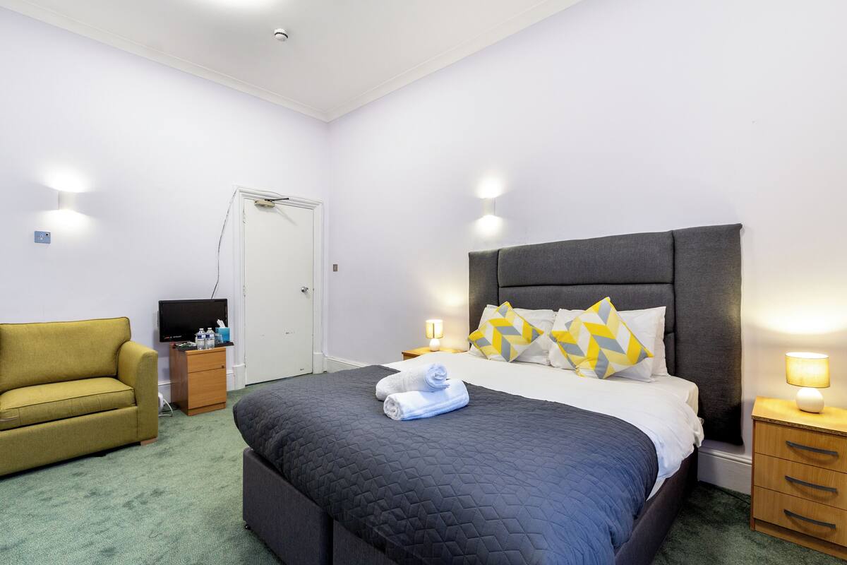 A hotel room with a bed, a sofa, a TV, and a bedside table with a lamp.