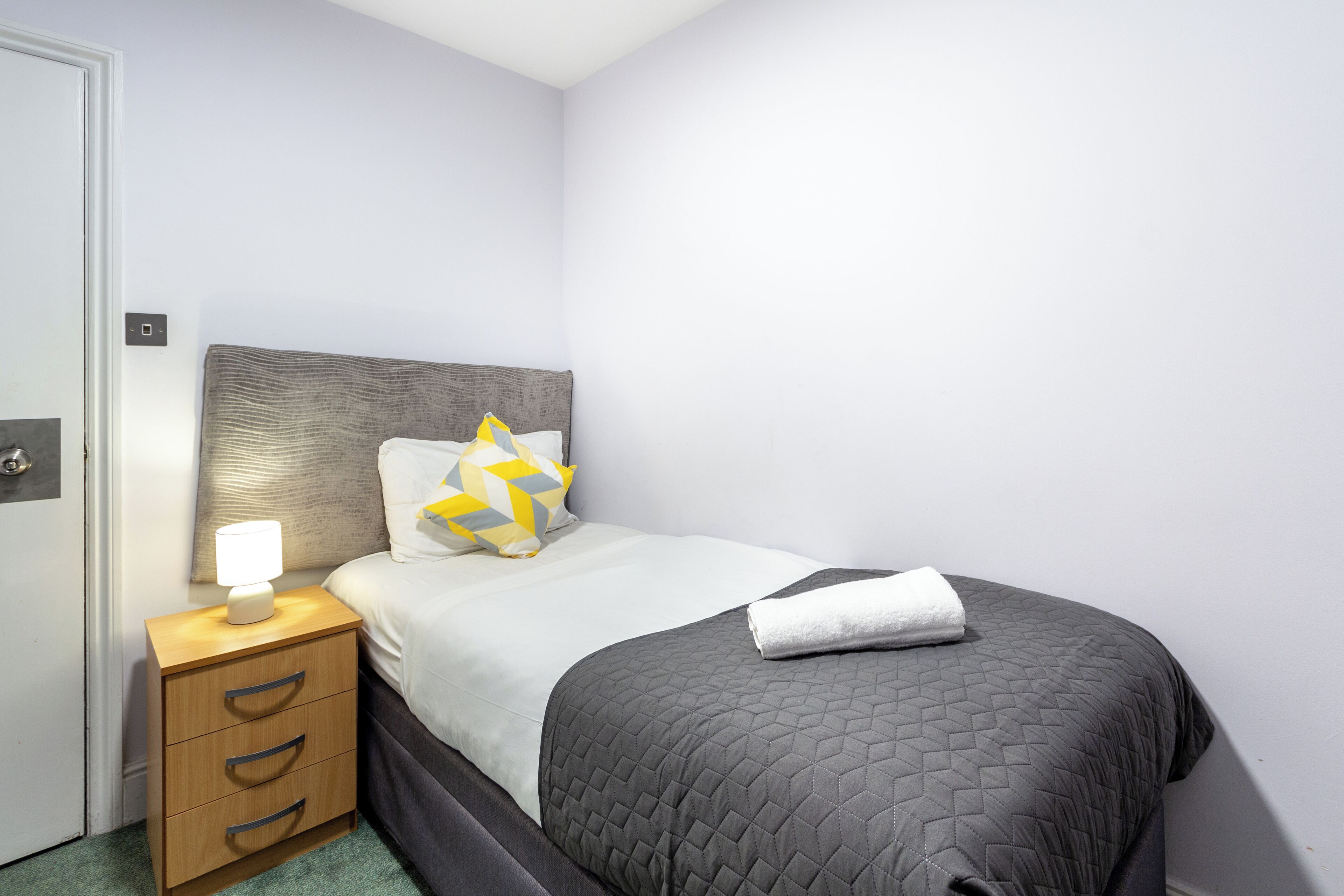 A neatly made bed with a grey quilt, a bedside lamp, and a nightstand with drawers.