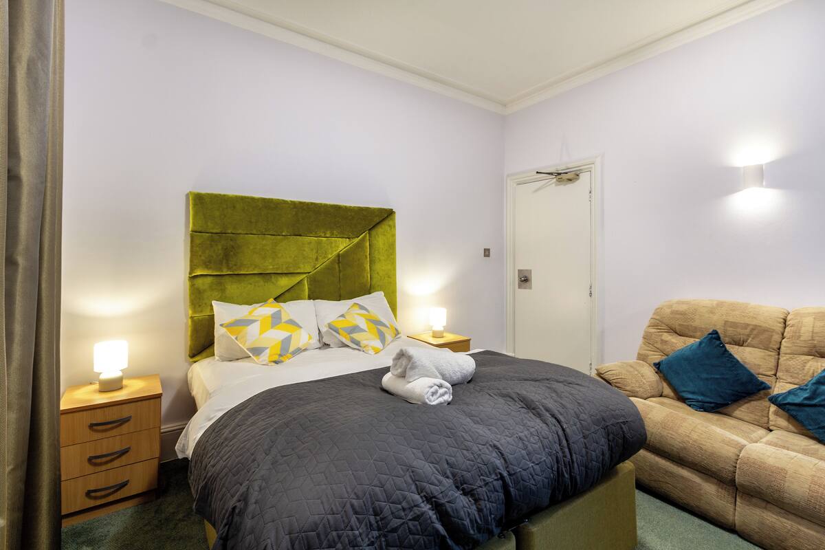 A neatly made bed with a green headboard, a bedside table with a lamp, and a couch with blue cushions.