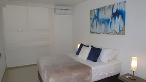 Quarto com cama, mesa de cabeceira, abajur e pintura abstrata.