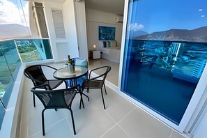 A balcony with a glass table and chairs, overlooking a pool and a mountainous landscape.