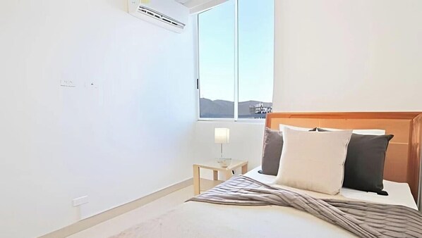 A bedroom with a bed, bedside table, lamp, and a window with a view. - Apartamentos Reserva del Mar - by SOHO (Santa Marta)