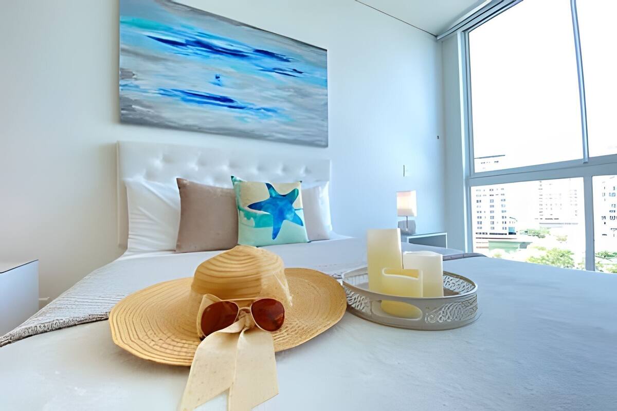 A hotel room with a bed, a painting of the ocean, a city view, a hat, sunglasses, and candles.