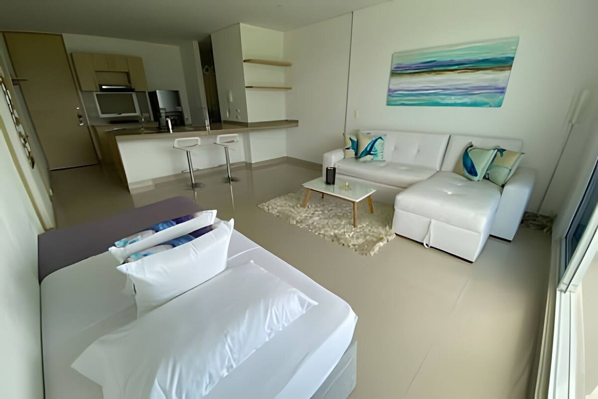 A modern living room with a sofa, a coffee table, and a TV. There is a kitchen area with a microwave and a refrigerator.
