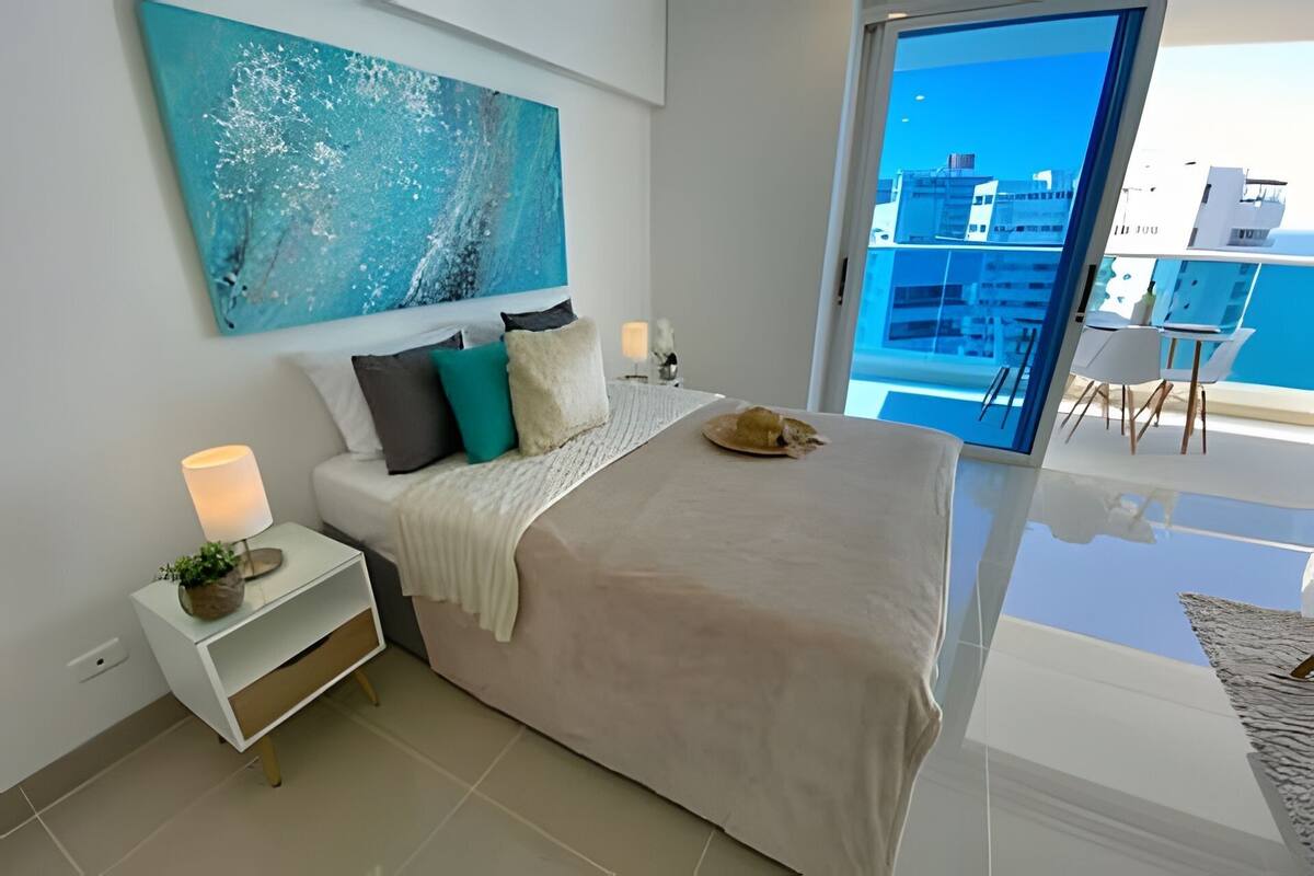 A modern bedroom with a large bed, a nightstand, a lamp, and a view of the city through a balcony.