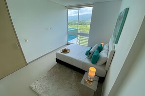 A modern bedroom with a bed, a small table with a lamp, and a view of a swimming pool and greenery through a large window. - Apartamentos Reserva del Mar - by SOHO (Santa Marta)