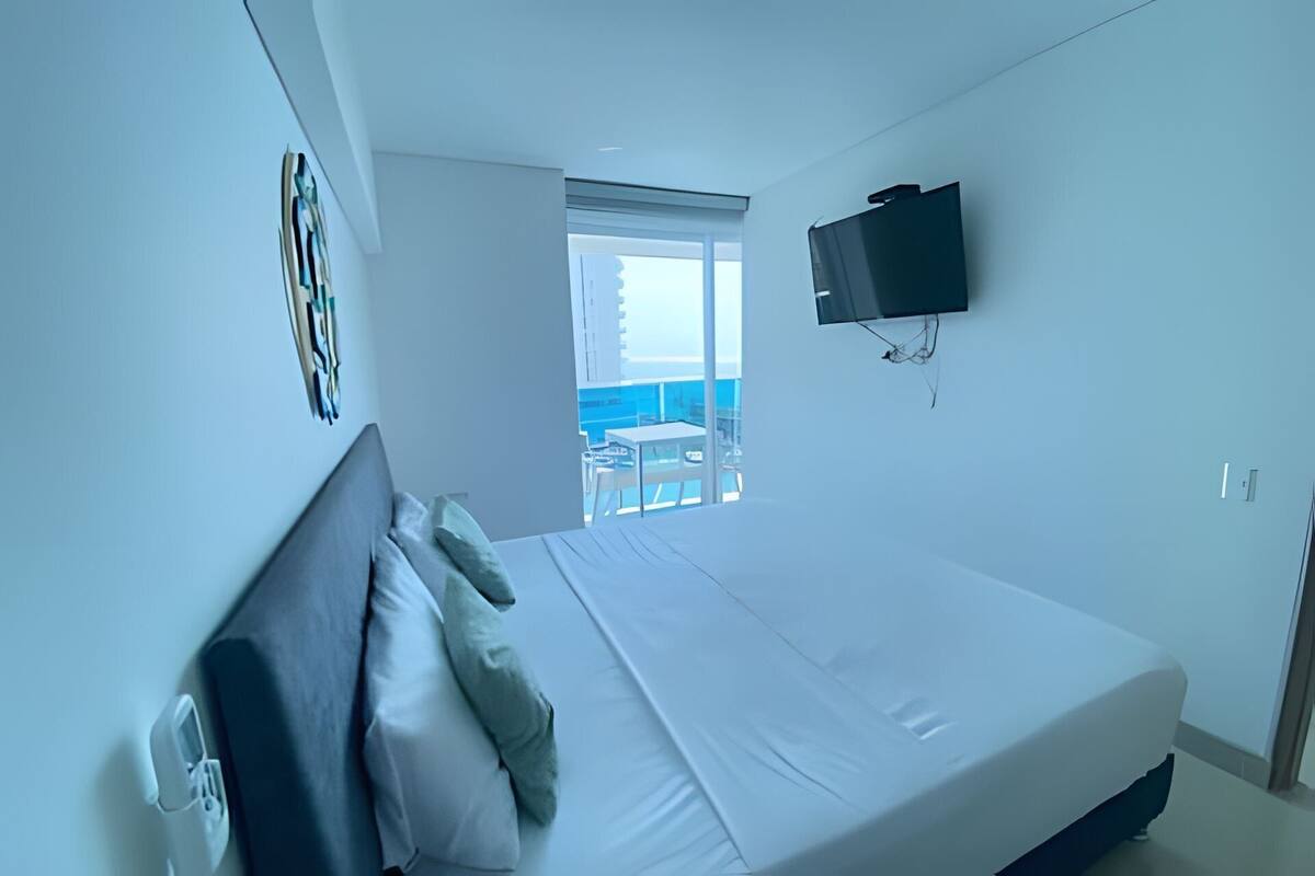 A modern hotel room with a large bed, a flat-screen TV mounted on the wall, and a balcony with a view of the sea.