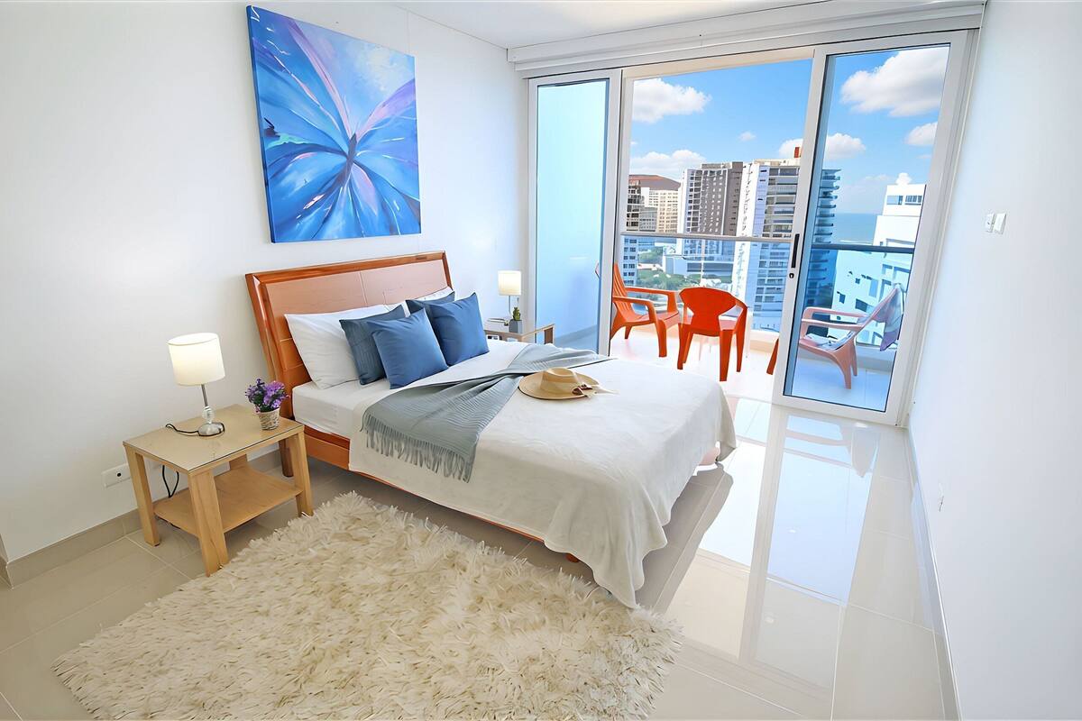A modern bedroom with a large bed, a nightstand, a fluffy rug, and a view of the city through a sliding glass door.