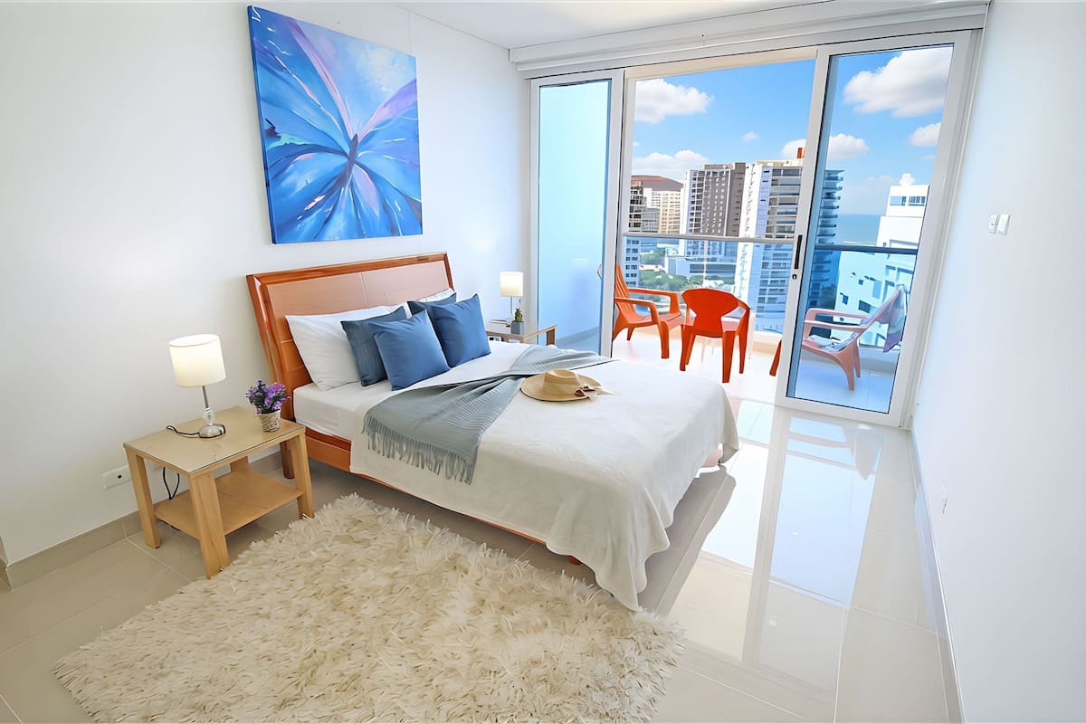 A modern bedroom with a large bed, a nightstand, a fluffy rug, and a view of the city through a sliding glass door.