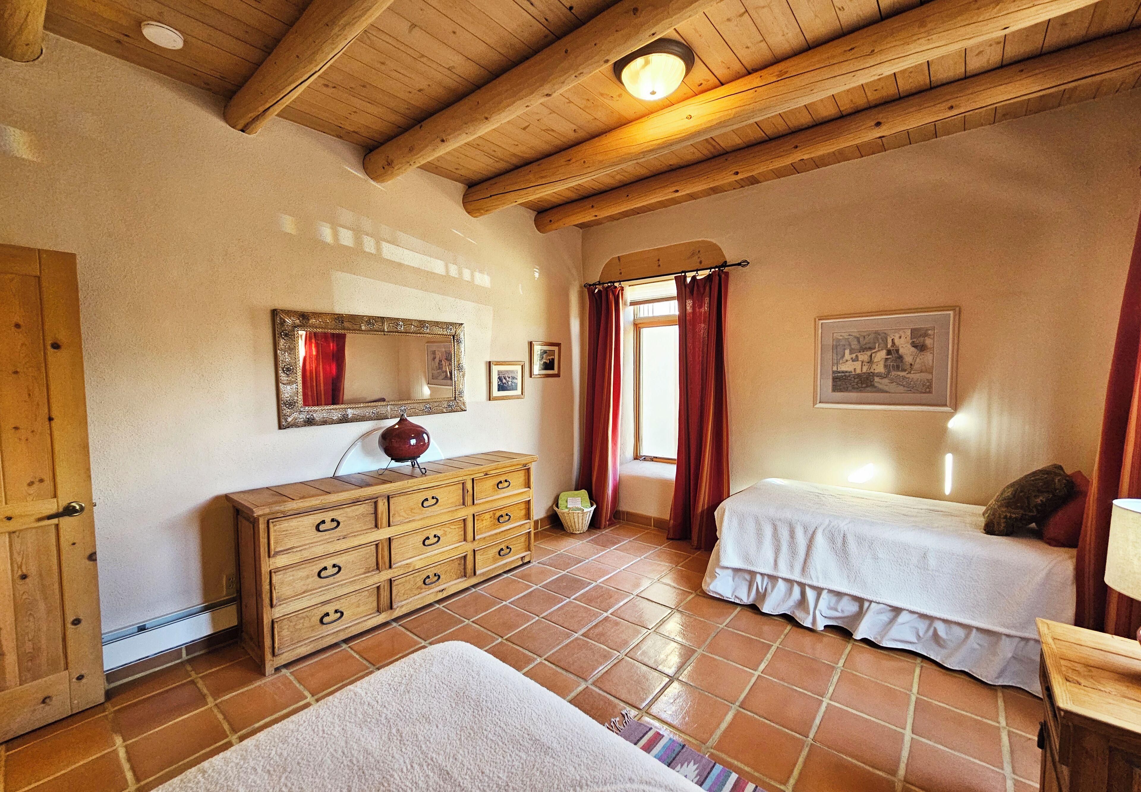A room with a wooden ceiling, a bed with a white bedspread, a wooden dresser with drawers, a mirror, and a window with red curtains.