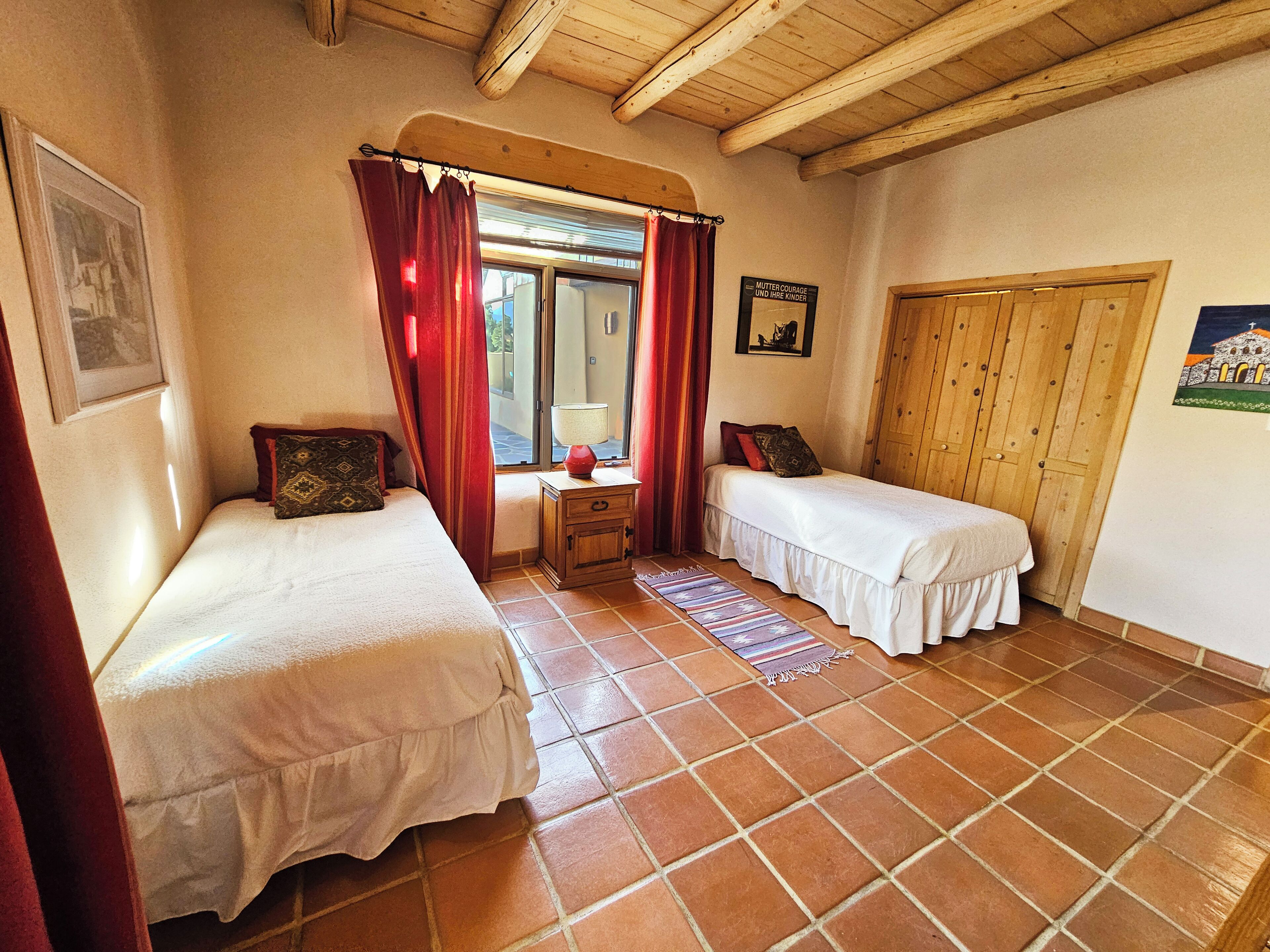 Una habitación con dos camas, un armario de madera, una mesita con lámpara y un ventanal con cortinas rojas.