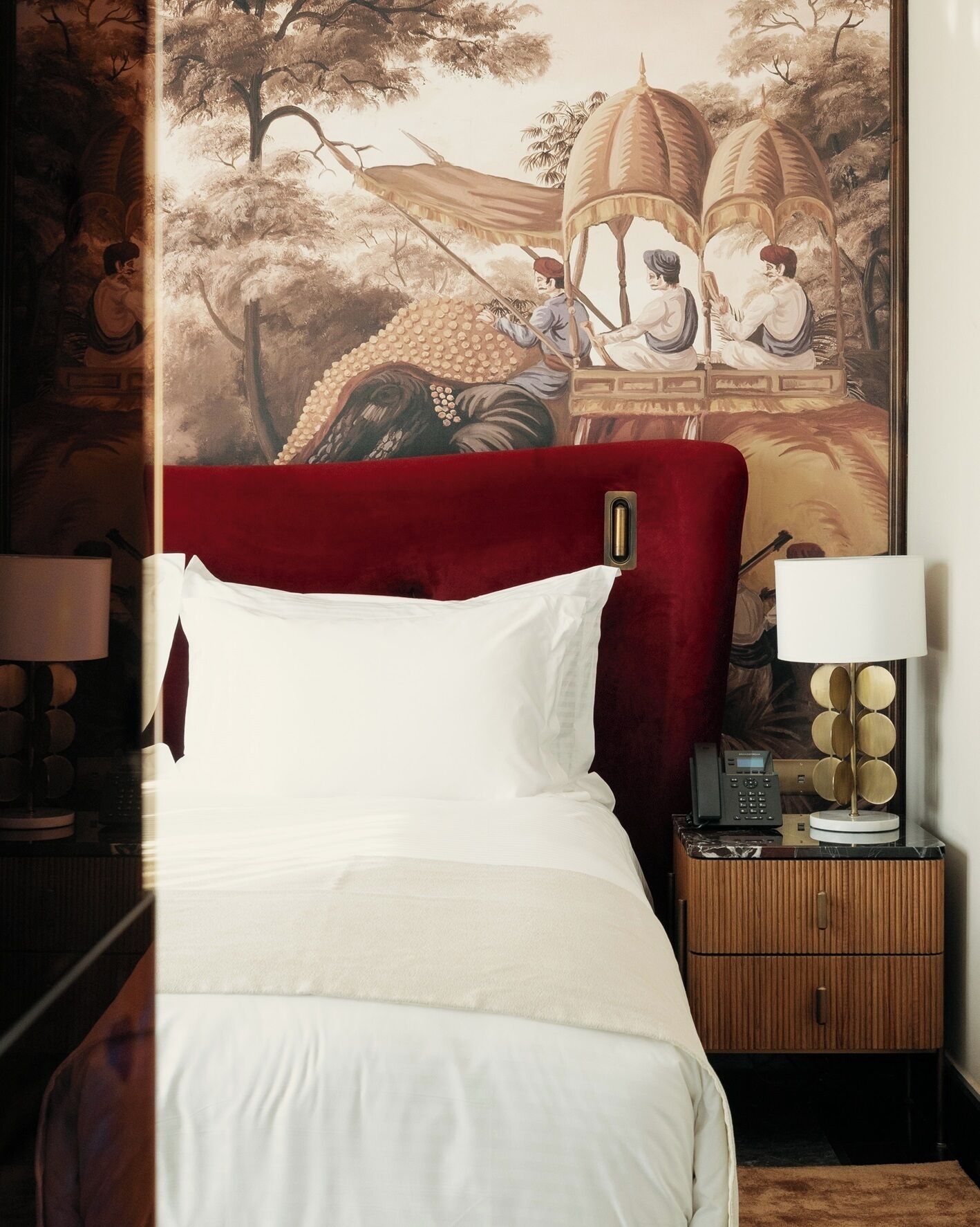 A hotel room with a large bed, a red headboard, a nightstand with a lamp, and a wall painting.
