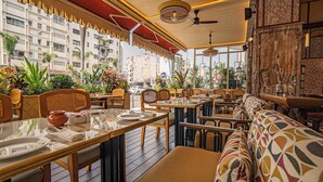 Un restaurante con terraza, mesas y sillas de madera, y vistas a edificios y plantas.