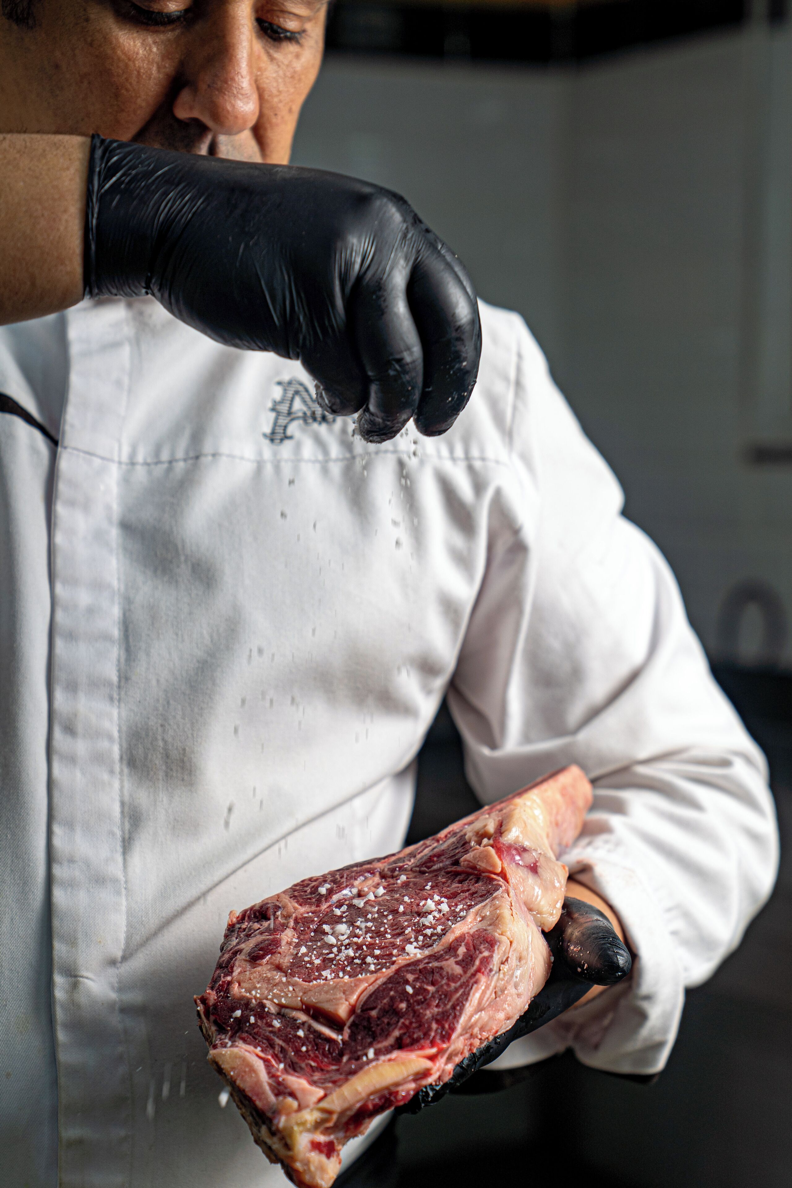 Una persona con un mandil blanco de chef sosteniendo un trozo grande de carne cruda con sal.