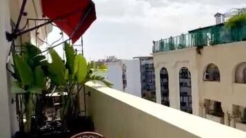 A balcony with a table set for dining, a red umbrella, and a view of buildings in the background.