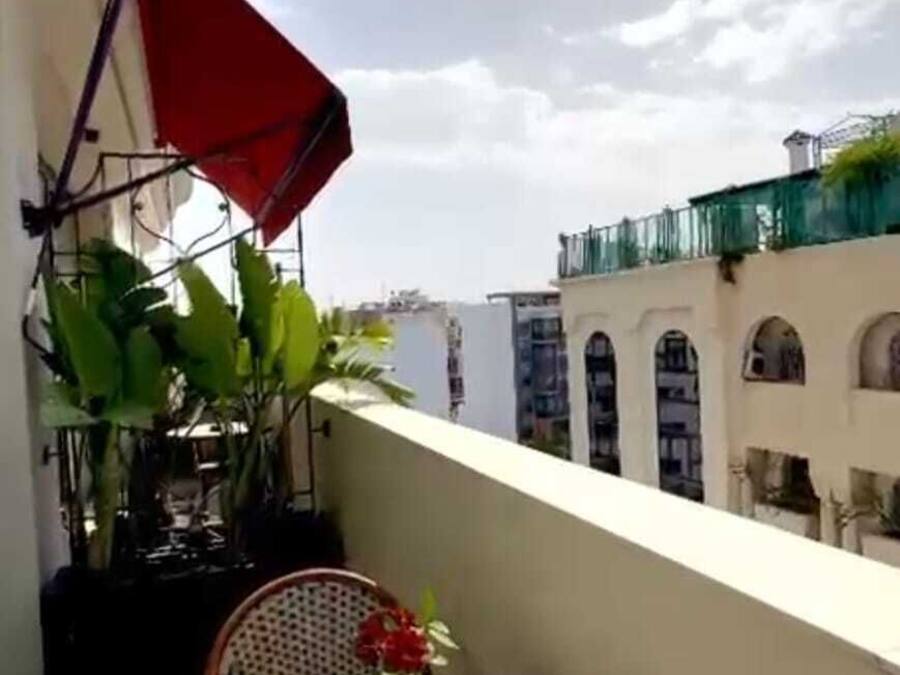 A balcony with a table set for dining, a red umbrella, and a view of buildings in the background.