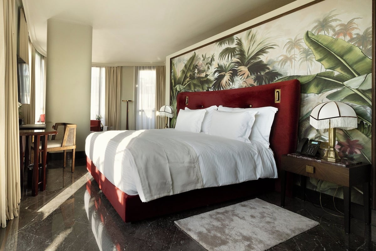 A hotel room with a large bed, a desk with a chair, a telephone, and a painting of tropical foliage on the wall.
