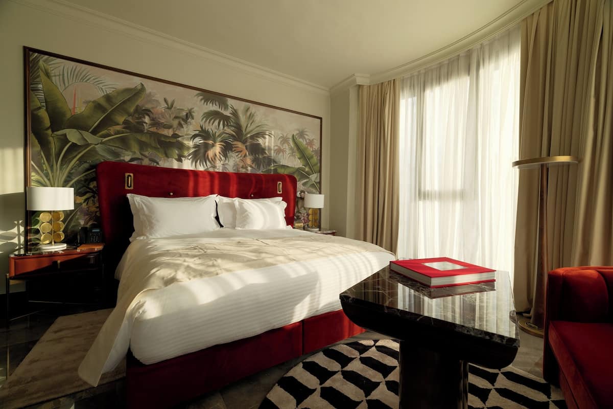 A hotel room with a large bed, a red headboard, a nightstand with a lamp, a red chair, a large painting on the wall, and a window with curtains.