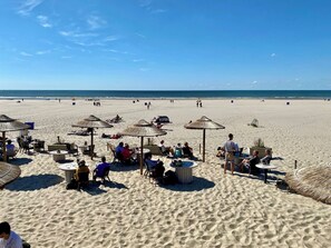A beach with thatched umbrellas and tables, people relaxing, and the ocean in the background.