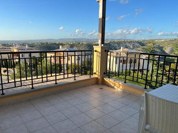 A balcony with a metal railing, a white chair, and a view of a residential area with houses and greenery.