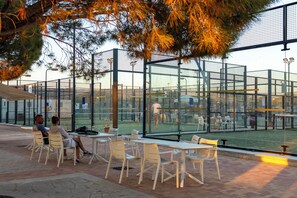 An outdoor seating area with tables and chairs, a glass enclosure, and a view of a tennis court. - Perfect Getaway for 2 With Shared Pool (Kouklia)