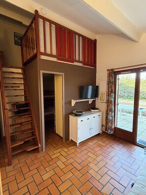 A room with a lofted sleeping area, a wooden ladder, a white cabinet with a television, and a door leading to an outdoor area.