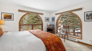 Un dormitorio con una cama grande, un escritorio y dos ventanas arqueadas que dan a vista de árboles.