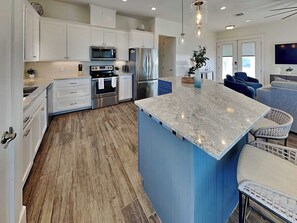 A modern kitchen with white cabinets, stainless steel appliances, a large island, and a blue accent wall. - Salty Sunrise | Bayfront Views + Private Pool (Rockport)