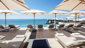 A poolside area with lounge chairs and umbrellas, overlooking a clear blue sea and a modern building.