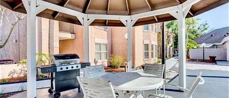 Un patio techado con una parrilla, mobiliario exterior y vista a un edificio residencial.