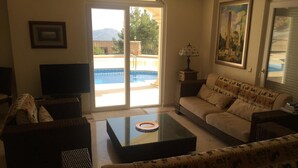 Un salon comprenant un canapé, des fauteuils, une table basse, un téléviseur et une grande fenêtre donnant sur une piscine.