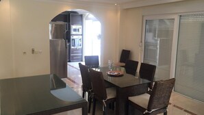 Un coin repas avec une table en verre et des chaises en osier, une cuisine en arrière-plan et une porte-fenêtre coulissante.